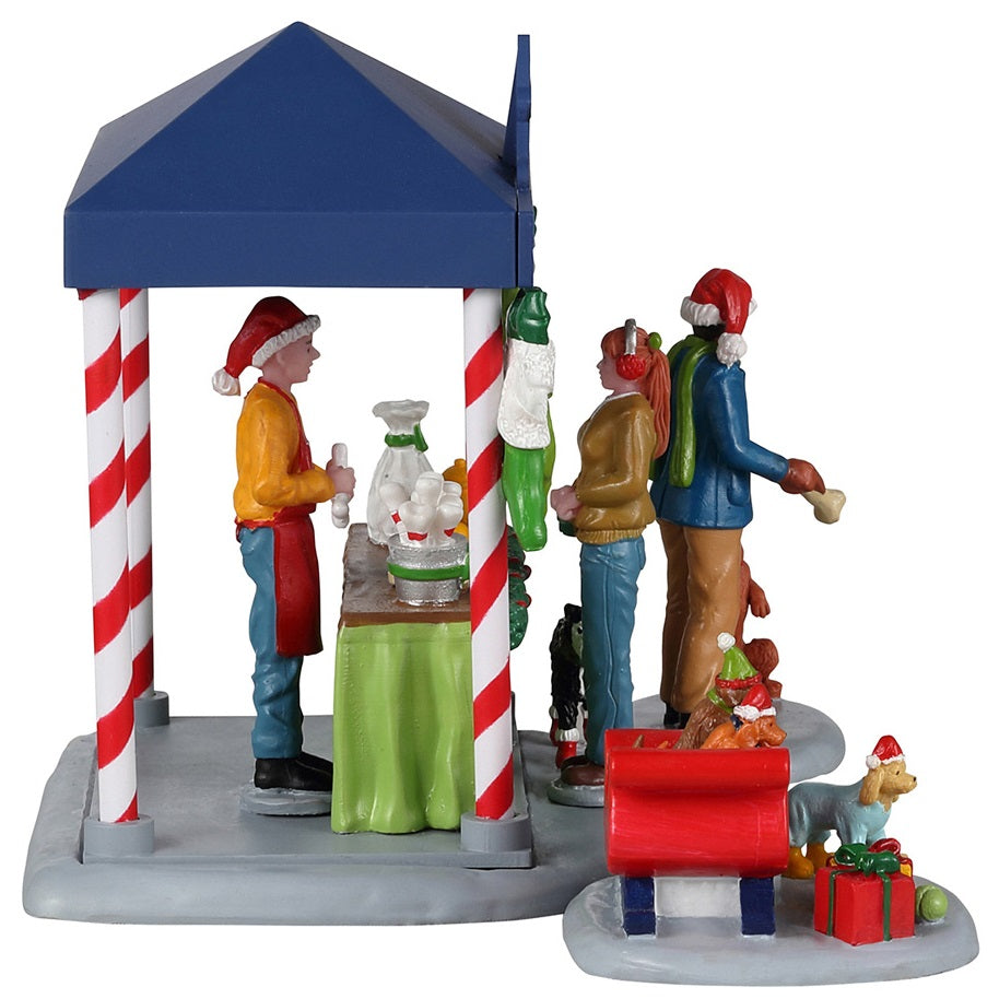 Resin table accent for a Lemax display. Designed to look like a booth that sells pet treats. It's decorated for Christmas. There's a woman handing her dog a bone while another is purchasing some food for her dog who is wearing red booties. There are two dogs wearing Santa hats in a red sleigh posing for pictures.