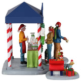 Resin table accent for a Lemax display. Designed to look like a booth that sells pet treats. It's decorated for Christmas. There's a woman handing her dog a bone while another is purchasing some food for her dog who is wearing red booties. There are two dogs wearing Santa hats in a red sleigh posing for pictures.