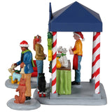 Resin table accent for a Lemax display. Designed to look like a booth that sells pet treats. It's decorated for Christmas. There's a woman handing her dog a bone while another is purchasing some food for her dog who is wearing red booties. There are two dogs wearing Santa hats in a red sleigh posing for pictures.