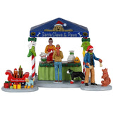 Resin table accent for a Lemax display. Designed to look like a booth that sells pet treats. It's decorated for Christmas. There's a woman handing her dog a bone while another is purchasing some food for her dog who is wearing red booties. There are two dogs wearing Santa hats in a red sleigh posing for pictures.