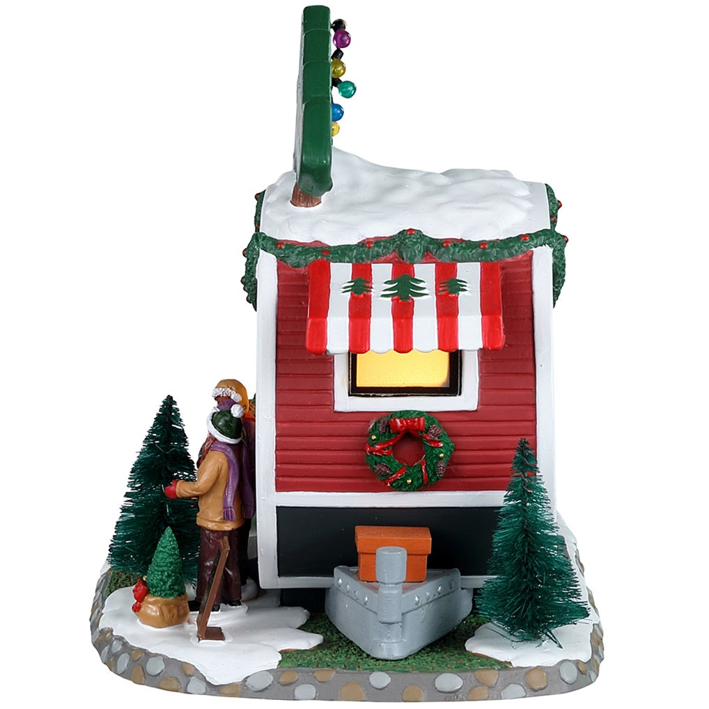 Porcelain lighted building for a Lemax display. Designed to look like a small red and white camper that's selling fresh Christmas trees. There are two people and a puppy checking out some of the trees outside.