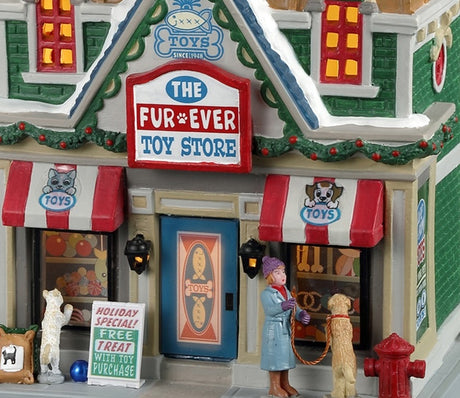 Porcelain lighted building for a Lemax display. Designed to look like a large toy store for pets. There's a white cat looking in the window on the left side of the building and a woman with a dog looking in the window on the right side of the building. There's snow on the roof and the sidewalk.