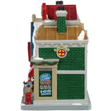 Porcelain lighted building for a Lemax display. Designed to look like a large toy store for pets. There's a white cat looking in the window on the left side of the building and a woman with a dog looking in the window on the right side of the building. There's snow on the roof and the sidewalk. There's a sign on the side that reads "Safe and fun toys for your pet! Choose from over 100 toys!"