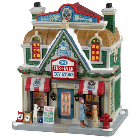 Porcelain lighted building for a Lemax display. Designed to look like a large toy store for pets. There's a white cat looking in the window on the left side of the building and a woman with a dog looking in the window on the right side of the building. There's snow on the roof and the sidewalk.