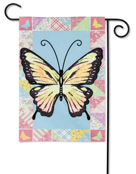 A flag featuring an Ornythion Swallowtail Butterfly. The background of this flag consists of different looking and different colored materials. Colors include: red, pink, yellow, light blue, lime green and orange