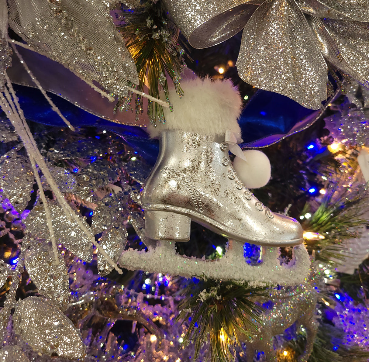 Silver ornament shaped like an ice skate. The cuff of the skate and the skate itself are furry and there are two pom poms tied to it.