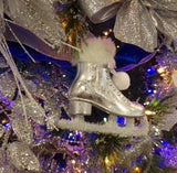 Silver ornament shaped like an ice skate. The cuff of the skate and the skate itself are furry and there are two pom poms tied to it.