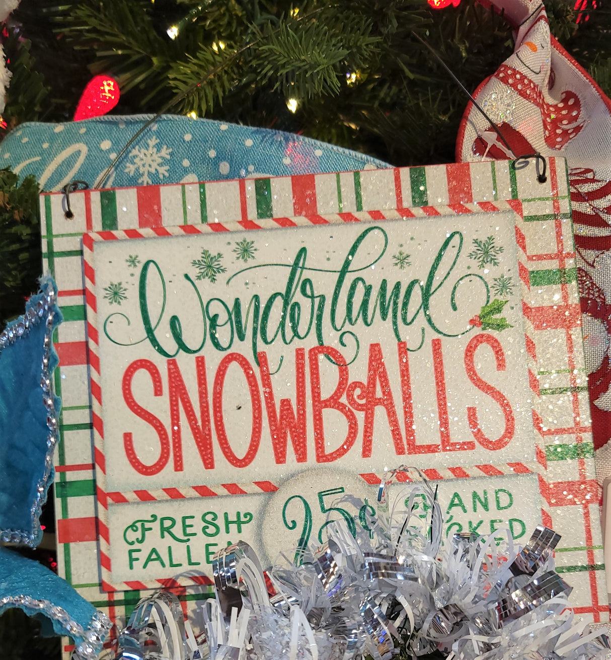 Sign hanging on a tree with other ornaments and tinsel. Sign reads "Wonderland Snowballs. Fresh Fallen. 25¢. Hand Packed."