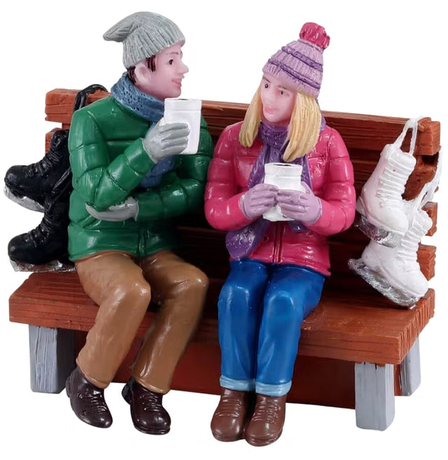 Polyresin figurine for a Lemax tabletop display. Designed to look like a man and a woman sitting on wooden bench. They're both wearing winter clothes and holding cups. There's a pair of black ice skates behind the man and a pair of white ice skates on the bench behind the woman