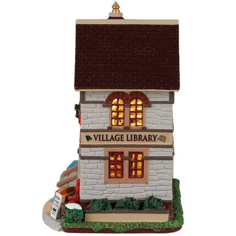 Porcelain lighted building for a Lemax display. Designed to look like a gray brick village library. There are red ribbon accents and decorated wreaths. Inside the window, you can see shelves of books and a table with chairs. There's a sign that reads "STORY TIME EVERY DAY AT 11 AM." There's a little snow on some of the bushes on the side.