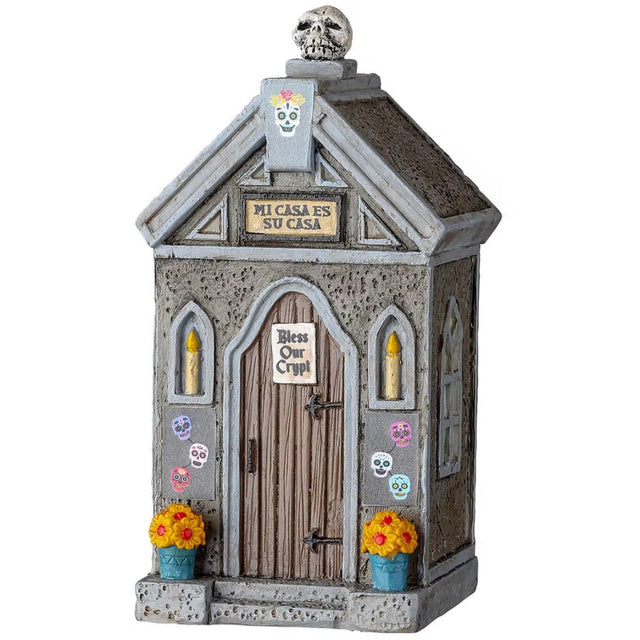 Resin village accessory for a Spooky Town Lemax display. Designed to look like a stone crypt with a wooden door. There are colorful skull accents and lit candles in the windows. The sign reads "MI CASA ES SU CASA." There's a sign on the door that reads "Bless our crypt." There are two planters with flowers on either side of the door.