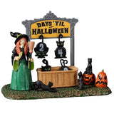 Resin village accent for a Spooky Town Lemax display. Designed to look like a witch holding a broom and a wand standing next to a Halloween countdown. The sign reads "Days 'Til Halloween" and there are removable numbers hanging off of it. There's a basket on the grass holding all the other numbers. There are black cats and jack-o-lantern accents. The number of days in this image are 24 days until Halloween.