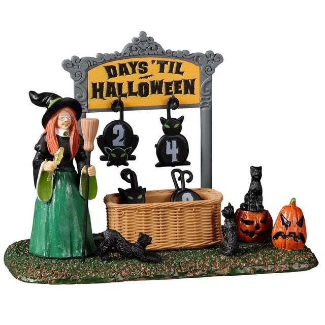 Resin village accent for a Spooky Town Lemax display. Designed to look like a witch holding a broom and a wand standing next to a Halloween countdown. The sign reads "Days 'Til Halloween" and there are removable numbers hanging off of it. There's a basket on the grass holding all the other numbers. There are black cats and jack-o-lantern accents. The number of days in this image are 24 days until Halloween.