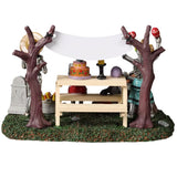 Resin village accent for a Spooky Town Lemax display. Designed to look like four skeletons enjoying a Day of the Dead celebration. There are tombstones, carved pumpkins and a banner hanging from the trees. There's a picnic table with a skull, a candle, a decorated cake and a rat on it.