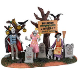 Resin village accent for a Lemax Spooky Town display. Designed to look like three skeletons wearing costumes in a graveyard. One skeleton is dressed like a vampire, one skeleton is dressed like a princess and the other skeleton is dressed like a witch and is petting a skeleton dog. The trees are decorated with pumpkin lights and there are black crows sitting on some branches.