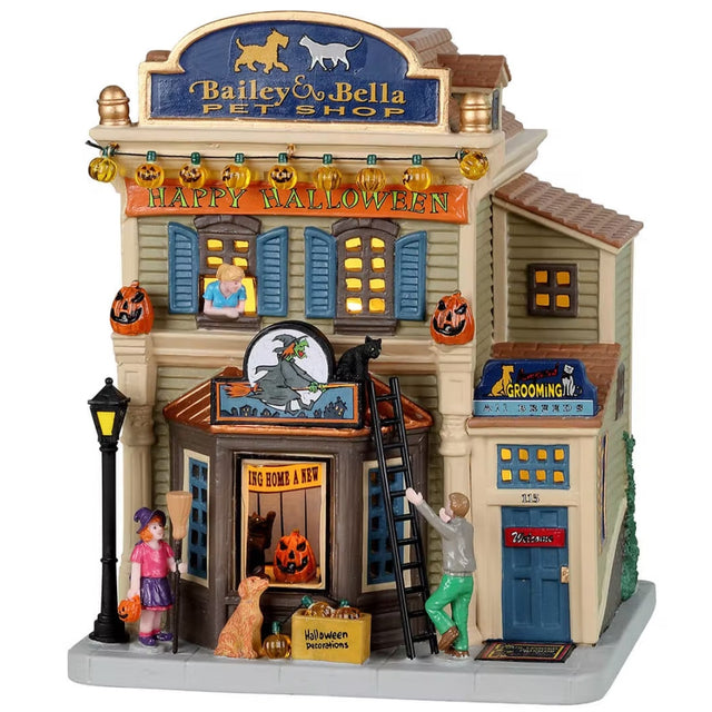 Porcelain lighted building for a Lemax display. Designed to look like a large pet shop. There are pumpkin lights hanging off the roof and a banner that reads "HAPPY HALLOWEEN!" There's a man putting a ladder against the building to get a black cat off the roof. There's a little girl dressed as a witch and a dog looking into the window of the building.
