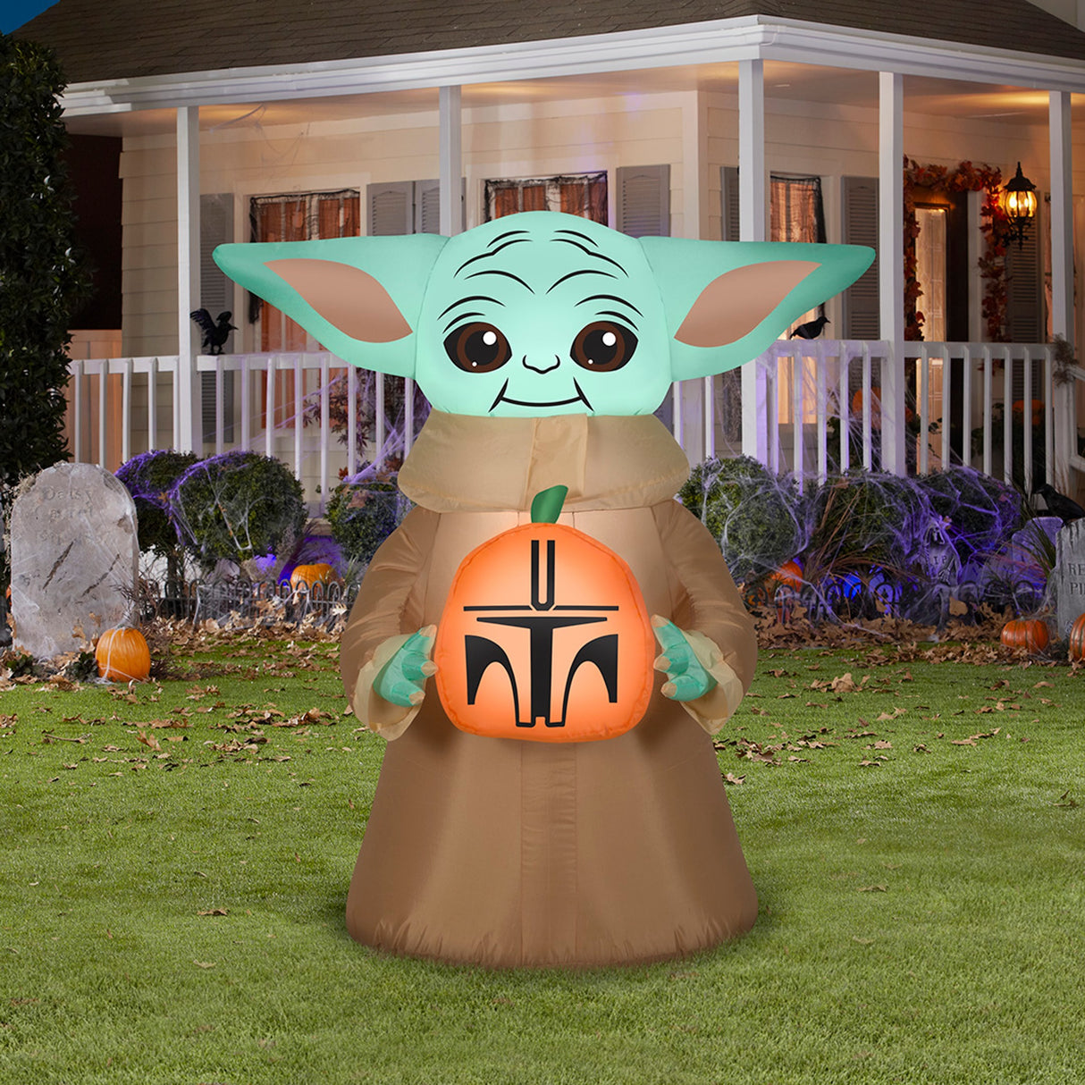 Halloween inflatable decoration shaped like baby Grogu from the show The Mandalorian. He's holding a small pumpkin that's been carved to look like a Mandalorian helmet.