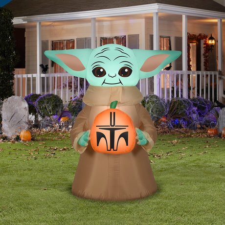 Halloween inflatable decoration shaped like baby Grogu from the show The Mandalorian. He's holding a small pumpkin that's been carved to look like a Mandalorian helmet.