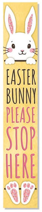 A vertical porch sign that reads "Easter Bunny Please Stop Here" on a yellow background. The top of a bunny is seen at the top and the bunny's feet are at the bottom
