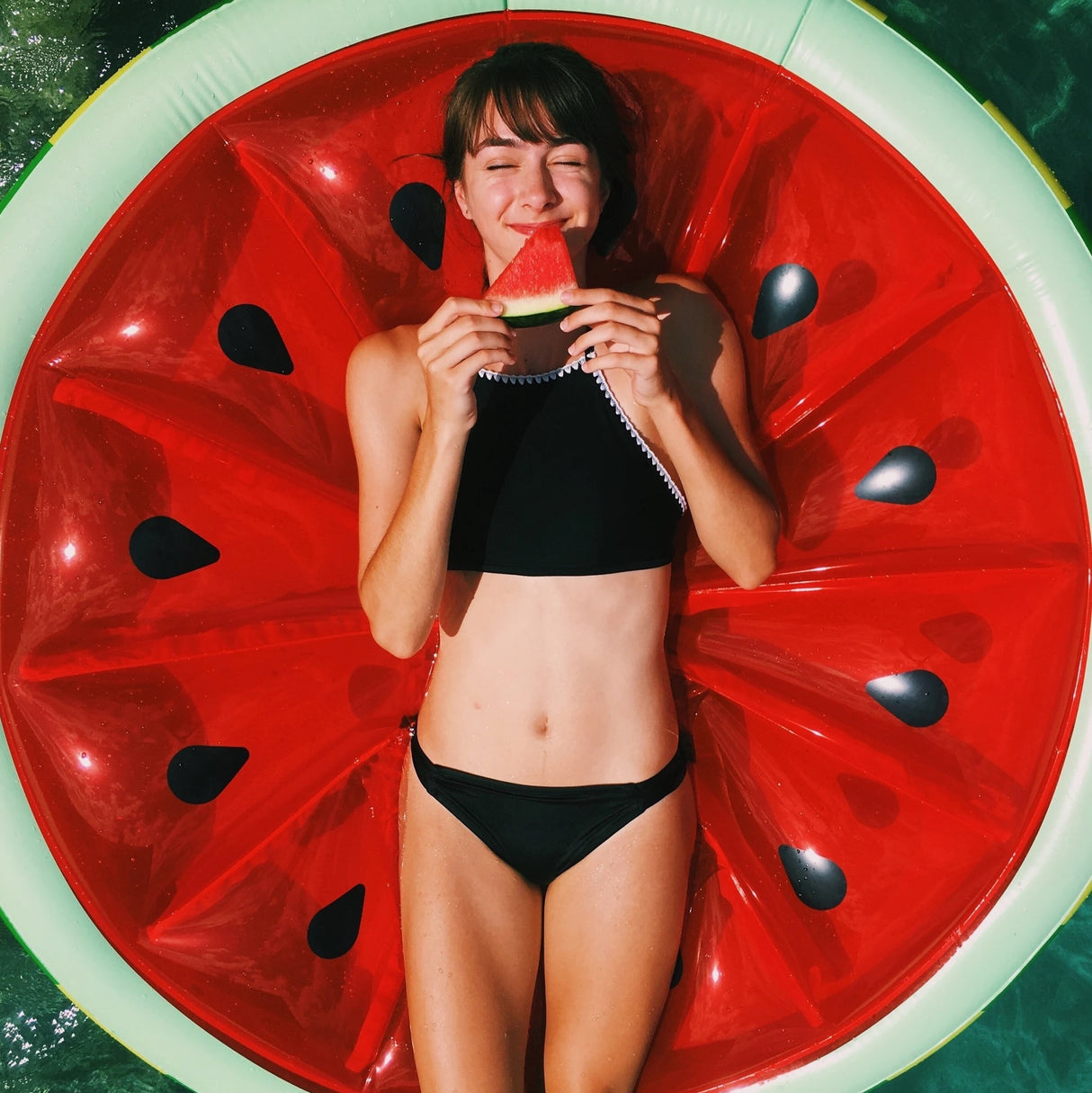 This is a lifestyle image showing a woman laying on the float while eating a piece of watermelon.