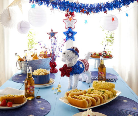 Patriotic tinsel tabletop decoration shaped like a dog holding a red star in its mouth. The dog is wearing a red, white and blue top hat and a red, white and blue jumper with a holographic star on the side.