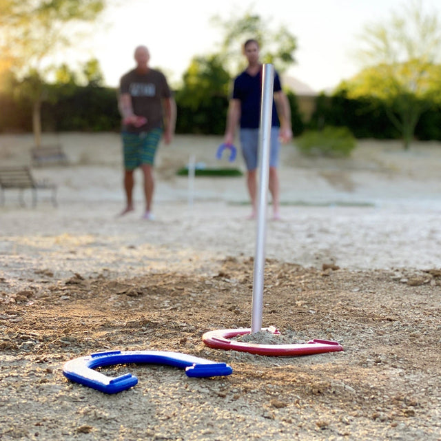 This is a lifestyle image showing one blue horseshoe and one red horseshoe next to one of the stakes. You can see two men in the background getting ready to throw more horseshoes.