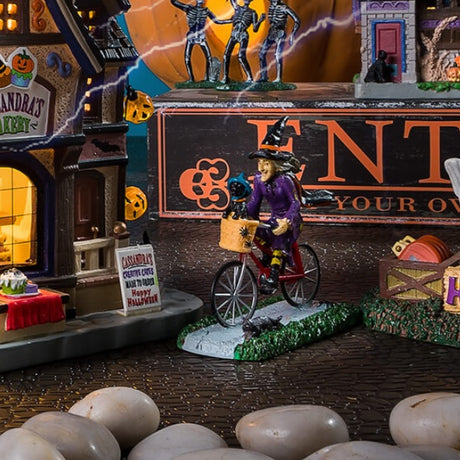 Polyresin figurine for a Lemax Spooky Town display. Designed to look like a witch on a bicycle. She's wearing a black witch hat, a black and orange cape and a purple dress. The bike is red and has a basket in the front with spiders on it. Inside the basket is a black cat wearing a blue witch hat and an orange collar.