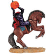 Polyresin figurine for a Lemax Spooky Town display. Designed to look like the Headless Horseman. The horse is brown with red eyes. The "head" of the man is a jack-o-lantern. The man is wearing a purple leather suit and a black cape with red inside.