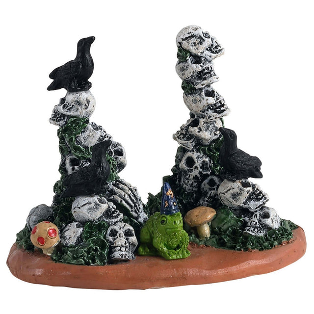 Polyresin village accessory for a Lemax display. Designed to look like a patch of dirt with two piles of skulls. There are crows and mushrooms and grass. In between the piles of skulls, there's a small green frog wearing a wizard hat.