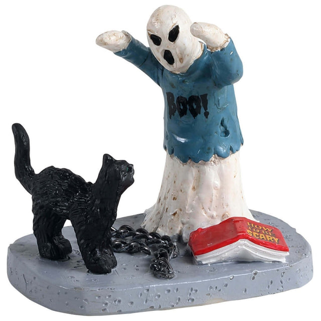Polyresin figurine for a Lemax Spooky Town display. Designed to look like a ghost wearing a tattered, blue sweater that reads "BOO!" There are chains, a black cat and a book on the ground. The red book cover reads "HOW TO BE SCARY!"
