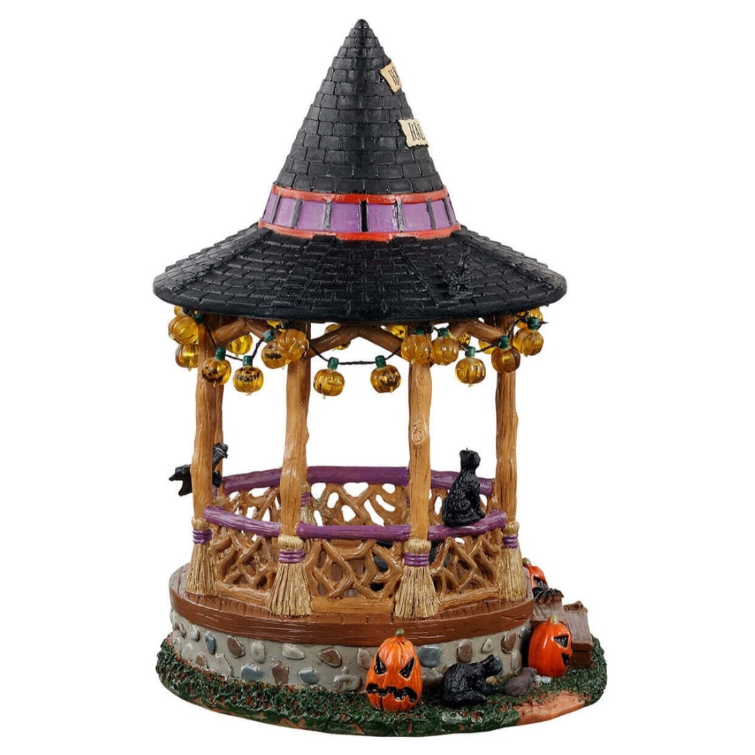 Polyresin village accent for a Lemax display. Designed to look like a wooden gazebo. The roof is shaped like a witch's hat and there's a sign that reads "HAPPY HALLOWEEN!" There are lights hanging from the roof that are shaped like jack-o-lanterns. There's a bat, a crow and a black cat. There's a cauldron with green potion inside. There are pumpkins around it and a toad. The columns of the gazebo are shaped like broomsticks.