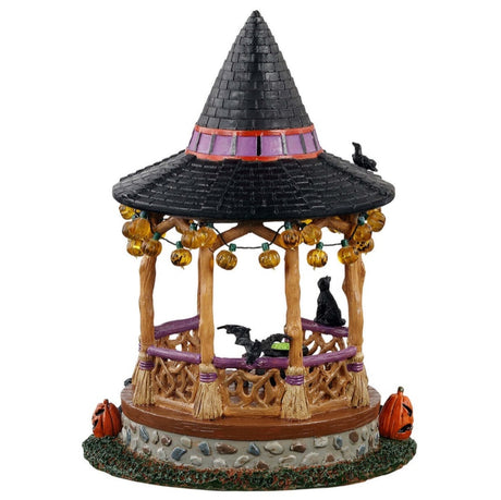 Polyresin village accent for a Lemax display. Designed to look like a wooden gazebo. The roof is shaped like a witch's hat and there's a sign that reads "HAPPY HALLOWEEN!" There are lights hanging from the roof that are shaped like jack-o-lanterns. There's a bat, a crow and a black cat. There's a cauldron with green potion inside. There are pumpkins around it and a toad. The columns of the gazebo are shaped like broomsticks.