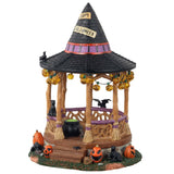 Polyresin village accent for a Lemax display. Designed to look like a wooden gazebo. The roof is shaped like a witch's hat and there's a sign that reads "HAPPY HALLOWEEN!" There are lights hanging from the roof that are shaped like jack-o-lanterns. There's a bat, a crow and a black cat. There's a cauldron with green potion inside. There are pumpkins around it and a toad. The columns of the gazebo are shaped like broomsticks.