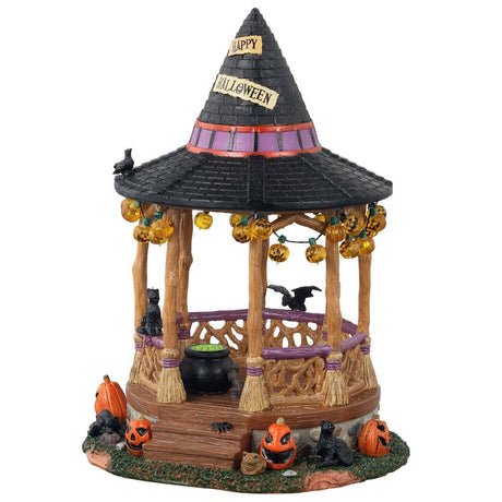 Polyresin village accent for a Lemax display. Designed to look like a wooden gazebo. The roof is shaped like a witch's hat and there's a sign that reads "HAPPY HALLOWEEN!" There are lights hanging from the roof that are shaped like jack-o-lanterns. There's a bat, a crow and a black cat. There's a cauldron with green potion inside. There are pumpkins around it and a toad. The columns of the gazebo are shaped like broomsticks.