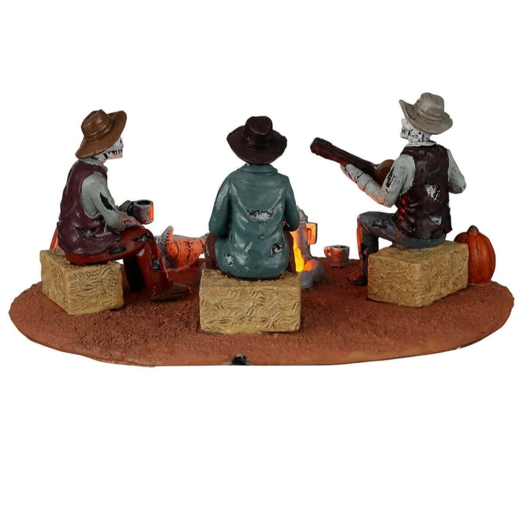 Plastic village accent for a Lemax display. Designed to look like three skeletons sitting around a campfire. They're sitting on bales of hay. There are carved pumpkins and an animal skull on the ground. All three skeletons are wearing cowboy hats and wearing ratty, old clothes. One skeleton is playing the guitar.