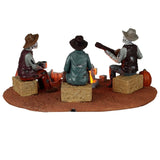 Plastic village accent for a Lemax display. Designed to look like three skeletons sitting around a campfire. They're sitting on bales of hay. There are carved pumpkins and an animal skull on the ground. All three skeletons are wearing cowboy hats and wearing ratty, old clothes. One skeleton is playing the guitar.