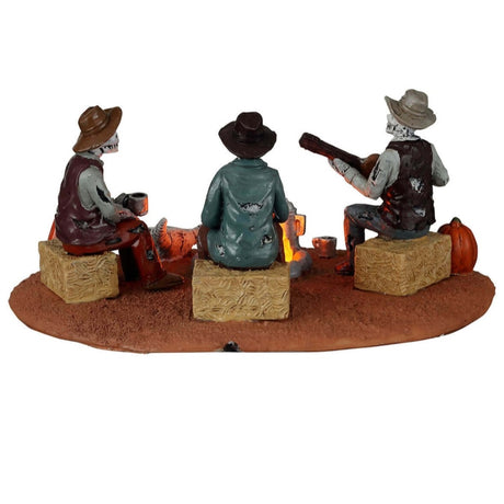Plastic village accent for a Lemax display. Designed to look like three skeletons sitting around a campfire. They're sitting on bales of hay. There are carved pumpkins and an animal skull on the ground. All three skeletons are wearing cowboy hats and wearing ratty, old clothes. One skeleton is playing the guitar.