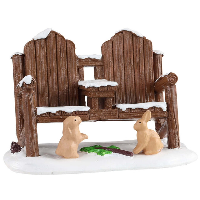 Polyresin figure for a Lemax display. Designed to look like a wooden bench with two seats. The bench is covered in snow and has two rabbits standing in front of it. There's a branch with some green leaves on it between the rabbits.