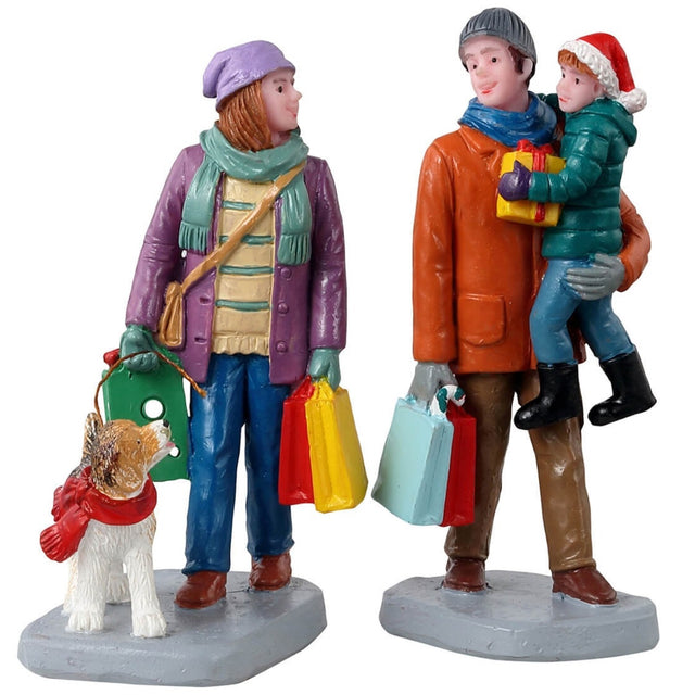 Polyresin figures for a Lemax Village tabletop display. A woman holding shopping bags and walking a dog. A man holding a small child and holding shopping bags. They're dressed for winter