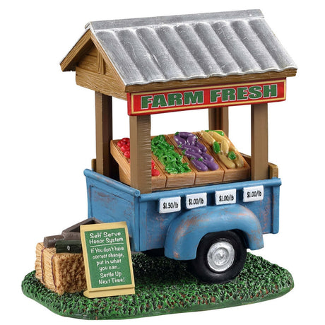 Polyresin figure for a Lemax display. Designed to look like a blue vegetable cart. There's a sign that reads "FARM FRESH" on the cart. Next to the cart, there's a sign on the ground that reads "Self serve honor system: If you don't have correct change, put in what you can... Settle up next time!" Inside the cart, there are tomatoes, green beans, eggplants and corn on the cob.