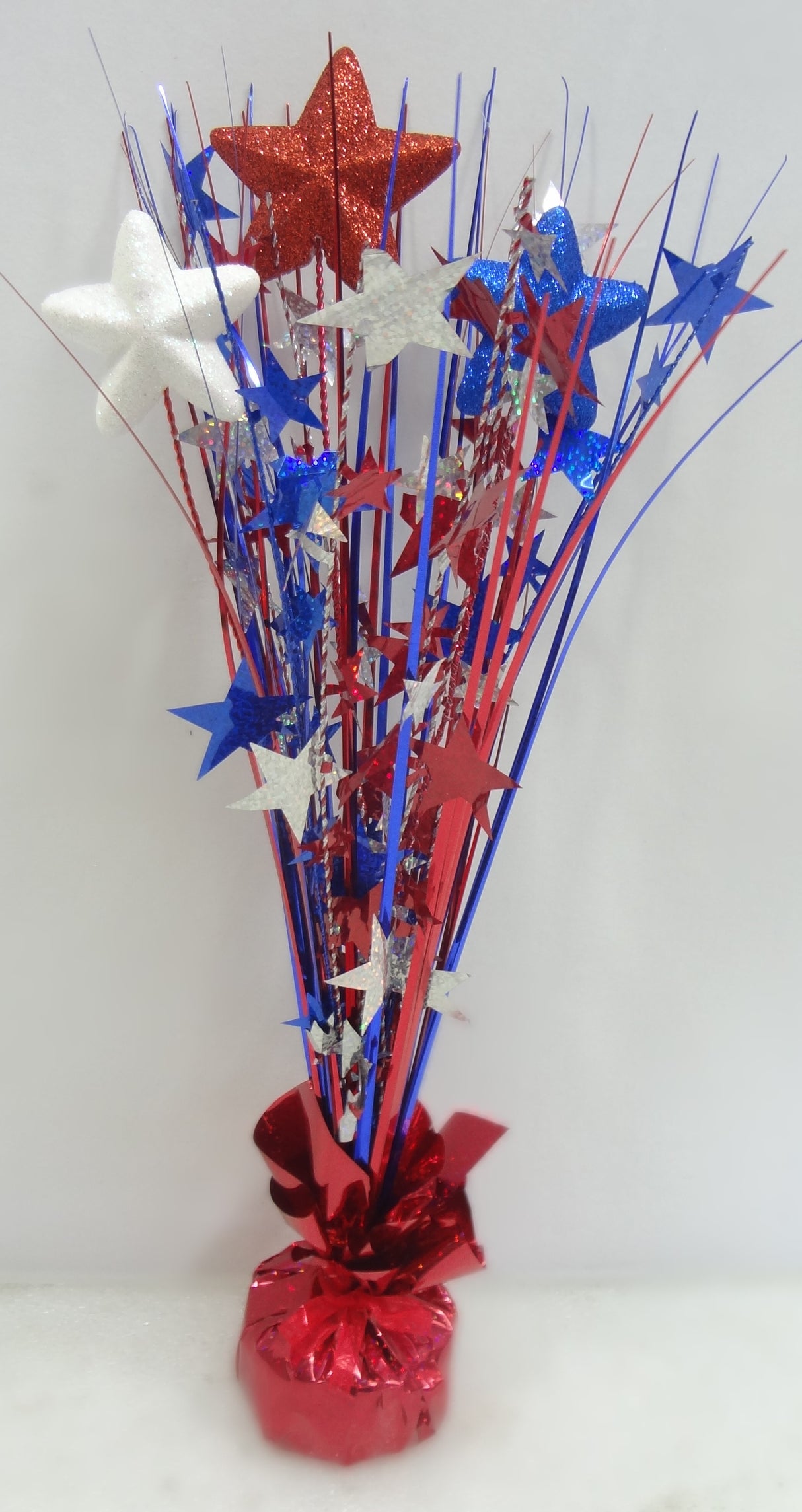 Patriotic tabletop decoration with a shiny, red tinsel base. Coming out of the base, there's red, white and blue tinsel with red, white and blue tinsel stars as well as foam stars with blue, red and white glitter.
