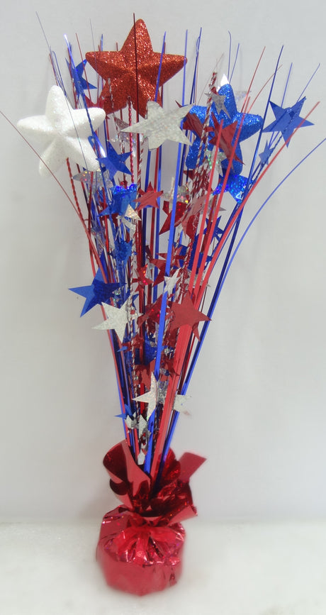 Patriotic tabletop decoration with a shiny, red tinsel base. Coming out of the base, there's red, white and blue tinsel with red, white and blue tinsel stars as well as foam stars with blue, red and white glitter.